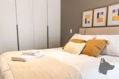 Dormitório com cama de casal, almofadas ocre, guarda-roupa e quadros na parede — Adore Canasvieiras em Canasvieiras, Florianópolis