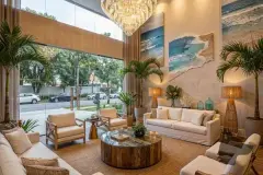 Hall de entrada do decorado com pé direito duplo, lustre de cristal, sofás brancos, plantas tropicais e quadros de praia na parede — Almare Jurerê em Jurerê, Florianópolis