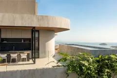 Foto real da varanda de cobertura com estrutura curva em concreto, cozinha gourmet integrada, balcão de madeira e vista panorâmica para o mar com ilhas ao fundo — Art Blue View em Jurerê, Florianópolis