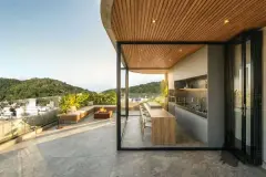 Foto real da varanda gourmet de cobertura com teto de ripas de madeira, bancada, lareira externa e vista para os morros e mar ao pôr do sol — Art Blue View em Jurerê, Florianópolis