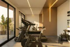 Perspectiva da academia compacta com esteira, bike spinning, TRX e rack de pesos, iluminação de teto em linhas douradas e vista para o jardim — Art Blue View em Jurerê, Florianópolis