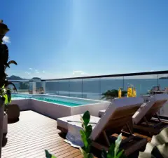 Perspectiva do rooftop com piscina de borda infinita, deck de madeira e vista para o mar de Jurerê, ângulo mais fechado — At One Jurerê Studios em Jurerê, Florianópolis