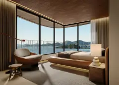 Perspectiva da suíte com janelas curvas do piso ao teto, cama de casal, poltrona orgânica e vista panorâmica para a Baía Norte e morros — Aura em Agronômica, Florianópolis