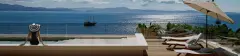 Perspectiva panorâmica da piscina de borda infinita com mulher de chapéu sentada à beira, deck de madeira, espreguiçadeiras, guarda-sol e veleiro histórico na Baía Norte de Cachoeira do Bom Jesus — Aurea Marina em Cachoeira do Bom Jesus, Florianópolis