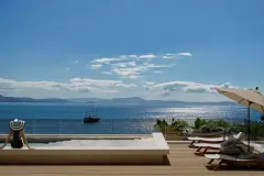 Perspectiva da piscina de borda infinita com mulher sentada à beira, deck de madeira, espreguiçadeiras, guarda-sol e veleiro na Baía Norte — Aurea Marina em Cachoeira do Bom Jesus, Florianópolis