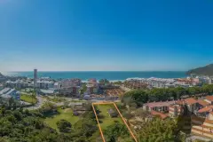 Foto aérea real do terreno do B7 Essence Brava delimitado em laranja com Praia Brava ao fundo e edificações vizinhas — B7 Essence Brava em Praia Brava, Florianópolis