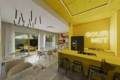 Espaço gourmet amarelo com bancada e cooktop, bancos industriais, letreiro neon Gourmet e mesas de jantar — BG Connect Canas em Canasvieiras, Florianópolis