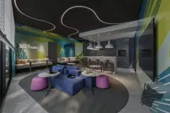 Espaço multiuso com sofá azul, pufes roxos, mesas bistrô, balcão bar e parede com grafite colorido — BG Connect Trindade 2 em Trindade, Florianópolis