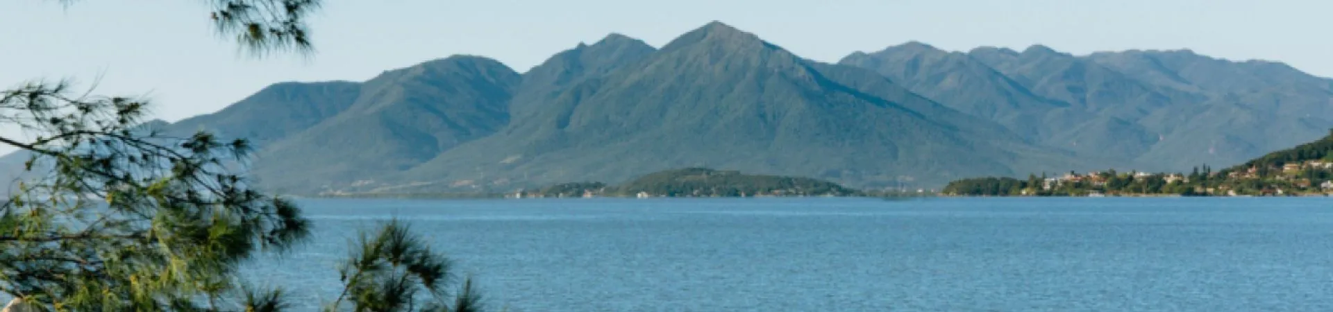 Vista do lago com montanhas ao fundo enquadrada entre galhos de pinheiro, entorno do D/Breeze Residence Club — D/Breeze Residence Club em Capoeiras, Florianópolis