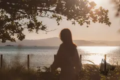 Foto real com silhueta de mulher observando pôr do sol sobre a Baía Norte enquadrada por árvores — D/Season Residence Club em João Paulo, Florianópolis