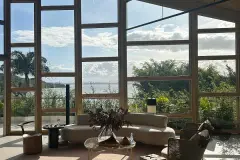 Foto real do lounge com sofá curvo bege, mesa de centro, plantas e janelão com vista para a Baía Norte e vegetação — D/Season Residence Club em João Paulo, Florianópolis