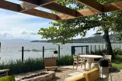 Foto real da área externa com pérgola em madeira, mesa redonda branca, jardim e vista para a Baía Norte com pedras — D/Season Residence Club em João Paulo, Florianópolis