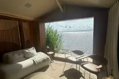 Foto real do lounge com sofá branco, plantas altas, janela com vista para a Baía Norte iluminada e painel de macramê na parede — D/Season Residence Club em João Paulo, Florianópolis