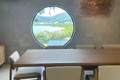 Foto real da sala de jantar com mesa retangular de madeira, janela circular com vista para a Baía Norte e luminária de palha rústica — D/Season Residence Club em João Paulo, Florianópolis