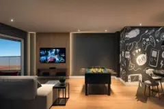 Perspectiva da sala de jogos do Essência Carlessi com pebolim, TV, sofá e parede com grafismos de games — Essência Carlessi em Córrego Grande, Florianópolis