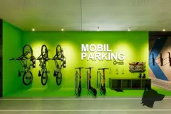 Bicicletário Mobil Parking com parede verde neon, bikes penduradas, patinetes elétricos e skates expostos — Imob Trindade On em Trindade, Florianópolis