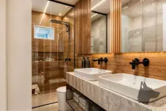 Banheiro com revestimento de madeira ripada, bancada de mármore com duas cubas e box de vidro — Infinity Residence em Ingleses, Florianópolis