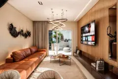 Sala de estar com sofá de couro caramelo, lustre circular, painel de TV em madeira e acesso à sacada — Infinity Residence em Ingleses, Florianópolis