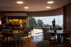 Perspectiva do salão de festas com mesas redondas, cozinha integrada e janelas para jardim — Lazuli Residence em Ponta das Canas, Florianópolis
