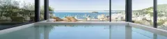 Piscina coberta com janelas panorâmicas e vista para o mar de Jurerê com barcos e cidade — Meraki Allure em Jurerê, Florianópolis