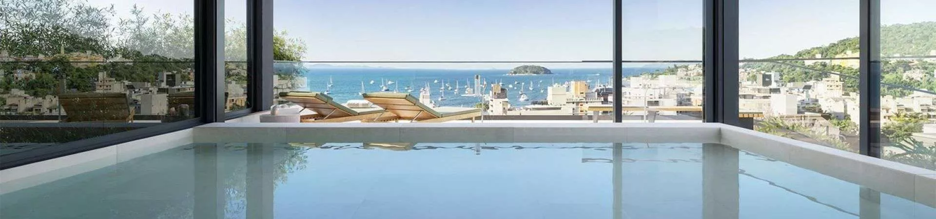 Piscina coberta com janelas panorâmicas e vista para o mar de Jurerê com barcos e cidade — Meraki Allure em Jurerê, Florianópolis