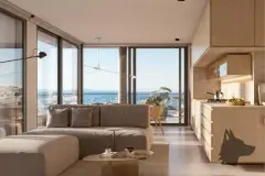 Sala de estar com sofá bege, cozinha integrada em madeira e janelas de canto com vista panorâmica para o mar — Meraki Allure em Jurerê, Florianópolis