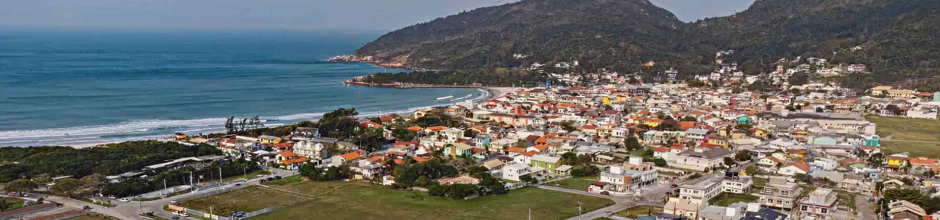 Vista aérea panorâmica da praia da Barra da Lagoa com mar azul-turquesa, casas coloridas e morro ao fundo em Florianópolis — Orla da Barra em Barra da Lagoa, Florianópolis