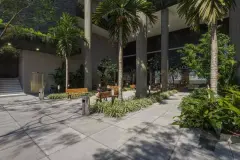Entrada do edifício durante o dia com palmeiras, bancos e fachada das lojas térreo — Saint Zambelli em Ingleses, Florianópolis