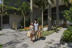 Casal caminhando na entrada do edifício com sacolas de compras, palmeiras e lojas ao fundo — Saint Zambelli em Ingleses, Florianópolis