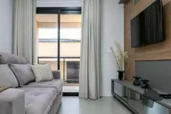 Sala de estar com sofá cinza, TV na parede, painel de madeira e porta de correr para varanda — Upside Beach em Ponta das Canas, Florianópolis