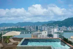 Piscina no rooftop com espreguiçadeiras, guarda-sol e vista ampla para o skyline de Florianópolis com montanhas ao fundo — VYU One em Coqueiros, Florianópolis