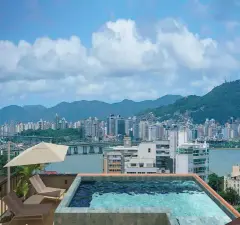 Piscina infinita no rooftop com espreguiçadeiras, mesa, guarda-sol e vista panorâmica para Florianópolis e a Ponte Hercílio Luz — VYU One em Coqueiros, Florianópolis