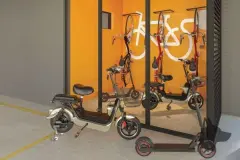 Bicicletário coberto com scooters elétricas, bikes suspensas e parede laranja com grafismo de bicicleta — VYU One em Coqueiros, Florianópolis