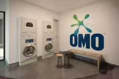 Lavanderia compartilhada com duas colunas de lava-seca, bancos e parede com logotipo OMO — VYU One em Coqueiros, Florianópolis